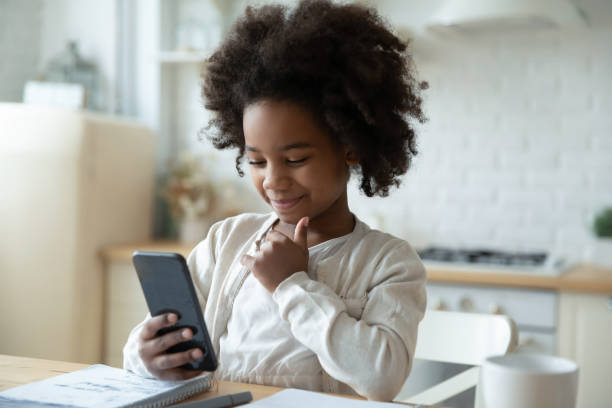 Smiling little African American girl watch distant class on smartphone, study online at home, happy small biracial kid use cellphone gadget, have web lesson on quarantine, homeschooling concept